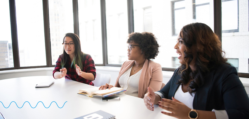 Three businesswomen seated on one side of a conference table in a sunny room